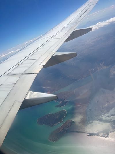 The image shows an aerial view from an airplane window, with the wing of the plane dominating the left side of the frame. Below the wing, a body of water with turquoise and darker blue hues is visible, dotted with several islands covered in vegetation. The land masses appear rugged and mountainous. In the distance, the terrain is hazy, with lighter patches suggesting clouds or atmospheric conditions. The sky is a clear, bright blue, with a few wispy clouds near the horizon. There are no people, animals, or man-made structures clearly identifiable on the islands or land. The scene depicts a natural landscape viewed from high above. The location in Eğirdir, Turkey, is not directly discernible from the visual information in the image itself.