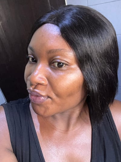 An adult female, appearing to be of dark skin tone, is depicted in a medium close-up, facing slightly left while looking towards the camera. She has short, straight, dark hair with a side part. Her lips are slightly parted, and her skin exhibits a glossy sheen. She is wearing a black sleeveless garment. The background is dark and indistinct, with a faint lighter area visible on the upper right and a dark vertical element on the left. The scene is located in Dzhidu, Nigeria.