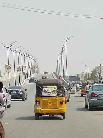 The image depicts a multi-lane elevated road or bridge in Gyadi Gyadi Arewa, Nigeria, under a hazy or overcast sky. In the immediate foreground, the back of a yellow three-wheeled motorized vehicle, commonly known as a Keke Napep or tricycle taxi, is centrally positioned, facing away from the viewer. This vehicle has a black canvas cover and a rectangular sign mounted on its rear, displaying the word "ASSAD" above an image of a bird or similar creature.
To the immediate left of the yellow tricycle, a silver sedan is partially visible, followed by a dark grey sedan (possibly a Honda model) with a visible green Nigerian license plate. On the right side of the road, further back from the yellow tricycle, a teal-colored sedan is present, with an individual in a light-colored shirt standing nearby.
The bridge itself features rows of evenly spaced streetlights equipped with solar panels along both sides, receding into the distance. Banners are attached to the lower portion of the streetlight poles on the left side of the road. The bridge barriers are concrete, topped with a wavy or corrugated pattern.
Further down the elevated roadway, two motorcycles carrying multiple riders are visible ascending the incline. In the mid-ground and background, other vehicles including cars, motorcycles, and another yellow tricycle are present on the road. Beyond the bridge structure on the right, several buildings and sparse vegetation are discernible, characteristic of an urban environment. The scene indicates active traffic flow on a clear day with noticeable atmospheric haze.