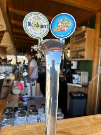 This medium close-up, portrait-oriented photograph, taken in Anatoliki Attiki, Greece, depicts two beer tap handles at what appears to be an outdoor bar or refreshment stand. The image is rotated counter-clockwise. The foreground is dominated by the gleaming chrome of a beer tap, reflecting a person (likely the photographer) and the sunny surroundings.

To the left of the chrome tap handle are two distinct tap knobs. The upper tap knob is blue and features a shield logo with red and white checks, along with partially legible text that includes "WANNUS HANISTER" and "ZYWOOLLA POAD" (or similar). The lower tap knob is gold-toned and clearly displays the "Warsteiner" brand name, a crown logo, the founding year "SEIT 1753" (since 1753), and the German phrase "DAS EINZIG WAHRE" (the only true one).

The background, partially out of focus, reveals a cluttered bar area constructed from wood. Various equipment, including what looks like an espresso machine, other bottles, and stacked plastic water bottles, can be seen. A person wearing a white shirt is partially visible behind the bar, tending to the setup. The bright lighting indicates it is daytime. The overall setting suggests a functional outdoor service area, possibly for an event, with a rustic aesthetic due to the wooden construction.