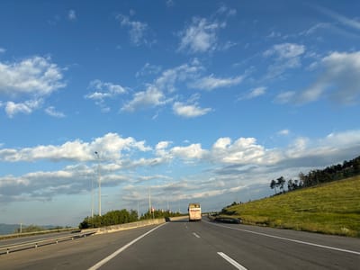 The image shows a highway in the vicinity of Tedotsminda, Georgia, under a blue sky with scattered white clouds. The road is paved with asphalt and has white lane markings. On the left side of the road, there is a concrete barrier and a metal guardrail. Beyond the guardrail, there are some green trees and vegetation. On the right side of the road, there is a grassy hill with scattered trees. In the distance, a white and pink bus is driving on the highway. There are several streetlights along the road. The overall scene depicts a rural highway in Georgia.