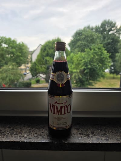 A tall, slender glass bottle of Vimto cordial is positioned centrally on a dark, speckled countertop. The bottle, filled with a deep red liquid, has a metallic cap and a label featuring gold, white, and red accents. The label has text in English and Arabic, including "VIMTO," "Cordial," "SINCE 1908," and "No Artificial Colours." Behind the bottle, a window offers a blurred view of green trees and a cloudy sky, suggesting an outdoor environment. The scene appears to be indoors, on a windowsill or ledge overlooking a residential area. The overall impression is one of a product placed for display or use, with the natural surroundings providing a soft, out-of-focus backdrop. It is not possible to determine the city of Oberhausen, Germany from the image alone.