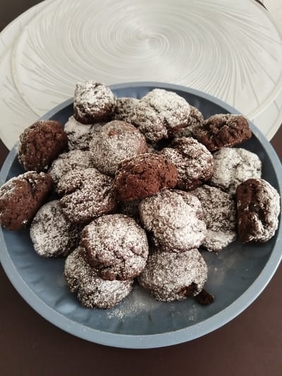 A close-up, overhead shot shows a pile of chocolate cookies dusted with powdered sugar on a blue plate. The cookies are round and craggy, with some appearing to be slightly broken. The plate is sitting on a dark brown surface, possibly a table. In the background, a white placemat with a swirled pattern is visible. No people or animals are present in the image. The scene appears to be indoors, likely in a kitchen or dining area. The location is stated to be Novostroika, Russia.
