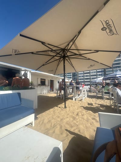 The image depicts a beach scene in Knokke-Heist, Belgium. A large Schweppes umbrella dominates the foreground, providing shade to a beach club. Several people are seen lounging on chairs and sunbeds. A white building likely belonging to the beach club is visible, with patrons dining and relaxing on the sand. The setting includes white furniture, sandy terrain, and a row of buildings in the background. The sky is blue, and the image is taken from a low angle, looking upwards towards the umbrella.
