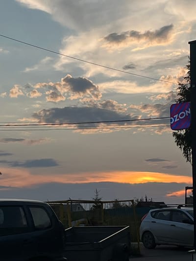 The image captures a dusky sky with scattered clouds illuminated by the setting sun, casting an orange glow on the horizon. In the foreground, a dark-colored SUV and a light-colored hatchback are parked. To the right of the vehicles, a trash bin and some yellow metal structures are visible. A sign with "OZON" written on it, illuminated with pink and blue stripes, is affixed to a pole on the right side of the image, near a tree. The scene appears to be on a street in Ludorvay, Russia, with a residential building or houses visible in the background. The overall atmosphere is calm, with the elements suggesting a typical evening scene in a semi-urban or rural setting.