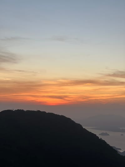 This media file displays a dramatic sunset over a mountainous landscape in El Cacao, El Salvador. The sky is a vibrant canvas of oranges, reds, and yellows, with wispy clouds stretched across the horizon. The sun, low in the sky, casts a warm glow that illuminates the clouds, creating a striking contrast with the dark silhouette of the mountains that dominate the right side of the frame. These mountains, covered in foliage, rise steeply, their jagged peaks outlined against the colorful sky. In the distance, several islands or rock formations rise from the hazy water, adding depth to the scene. The overall mood is one of tranquility and natural beauty, enhanced by the spectacular display of colors. The time of day is clearly sunset, and the weather appears clear, allowing for an unobstructed view of the celestial phenomenon. There is no visible text in the image.