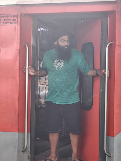 A man with a beard and turban stands in the doorway of a train car. He is wearing a teal t-shirt with a white graphic and dark blue shorts. He is holding onto the train door handles on either side of the doorway. The train car is red with a gray lower section. A sign on the exterior of the train car reads "बैठने/सोने के लिए 80 TO SEAT/SLEEP." This image was taken at the Itarsi Junction railway station in Itarsi, India.