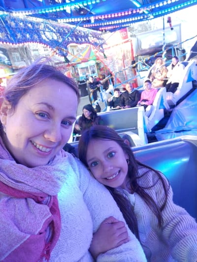 The image depicts a selfie taken from inside a brightly lit fairground ride, presumably in Aubagne, France. In the immediate foreground, a woman with light brown hair, wearing a white knitted sweater and a pink scarf, smiles directly at the camera. Adjacent to her, on her right, a young girl with long dark hair, wearing a light-colored knitted cardigan, leans against the woman's arm and also smiles broadly at the camera.

Both individuals are seated within a blue and white patterned ride compartment, which is illuminated by a cool blue light. The upper structure of the ride is visible overhead, adorned with numerous blue and pink string lights. In the middle ground, other individuals are seated in similar compartments of the same or an adjacent ride; these include a person in a pink hoodie, an older man in a dark jacket, and other adults. Background elements include other fairground attractions, such as a colorful stall with a striped roof, part of a larger white structure, and additional decorative lighting. A red and white circular emblem is partially visible in the distant background. The scene suggests an active fairground environment.