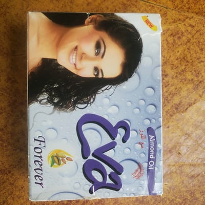 This is a media file of a product box, likely a cosmetic or personal care item, placed on a textured surface that appears to be a wooden table with a brown and orange mottled pattern. The box is positioned in the center of the frame.

The front of the box features a prominent image of a woman with dark hair and makeup, smiling directly at the viewer. Her face is well-lit, highlighting her features. The background of the box is a light blue with a pattern of water droplets.

Text on the box includes "NEW" in a yellow burst on the top left corner. Above the woman's head, on a purple band, is the text "Almond Oil" in English, and below that, in red Arabic script, the word "اللو" which translates to "al-luz" or "almond." In the center of the box, in large purple stylized lettering, is the brand name "Eva." Below the brand name, there is an illustration of a yellow teardrop shape containing almonds. At the bottom of the box, the word "Forever" is written in purple, with a metallic sheen.

The image conveys a sense of freshness and beauty, likely intended for skincare or haircare products. The setting is indoors, as indicated by the consistent lighting and the presence of the product box. There are no visible cues to suggest a specific time of day or weather conditions. The overall impression is that of a product advertisement.