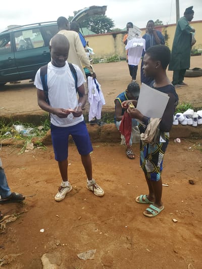 In Bahit, Nigeria, a group of people are gathered around a teal minivan with its trunk open. In the foreground, a young Black man wearing a white t-shirt, blue shorts, and white sneakers is looking down at some cash in his hands. He has a blue backpack on. To his right, a young Black woman wearing a dark top and a colorful patterned skirt is holding a white folder and a red cloth. Behind them, another Black man in a blue and yellow shirt and a black cap is kneeling, and several other people are visible in the background, including one wearing a dark green robe and a man in a light-colored robe. Several pairs of white sneakers are neatly arranged on the ground to the right of the woman. The ground is a mixture of dirt and debris, with some overgrown vegetation visible along a low wall in the background. The sky is overcast.