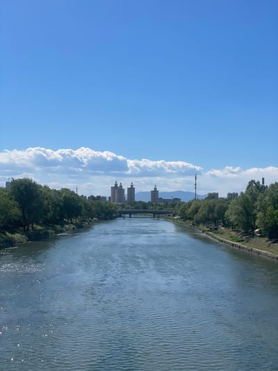 The image shows a river in the Changping District, China. The water is rippled, suggesting a slight breeze. Trees and vegetation line the riverbank. In the distance, several buildings are visible, including taller structures that could be residential or commercial towers. A communications tower is visible behind the buildings, further away, with mountains also in the distance. The sky is bright blue with puffy white clouds.