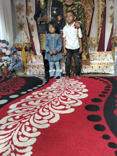 The image depicts a formal indoor gathering, likely a wedding celebration, set in Cottrueng, Indonesia. A family, including two children, stands on a red carpet with a decorative white and black pattern. The children, a girl in a denim dress and a boy in a white polo shirt and dark pants, are positioned in the foreground. Behind them, adults in traditional attire, one in a dark green and gold patterned garment and another in a dark formal jacket with gold embellishments, stand partially visible. The background features ornate drapes and decorative elements, suggesting a ceremonial setting. The lighting is consistent with indoor ambient light, casting soft shadows.