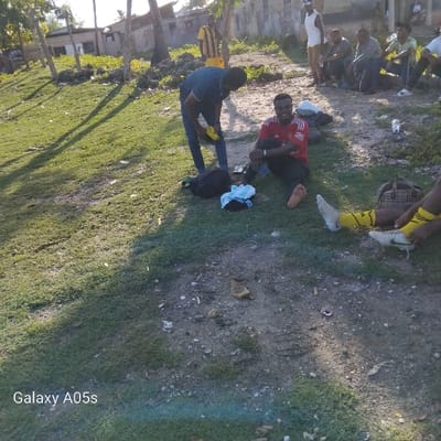 The image shows a group of men in Nungwi, Tanzania, near the coordinates -5.726 latitude and 39.301 longitude. The men are standing and sitting on a grassy area. A man in a red shirt is sitting on the ground, with his legs outstretched. He is wearing yellow and black socks and black shoes. Another man is standing nearby and appears to be tying his shoes. There are bags and other objects scattered around the men. The environment is outdoor and appears to be in the shade. There are some trees in the background. The image was taken by a phone. A logo for a Samsung Galaxy A05s phone can be seen at the bottom of the image.