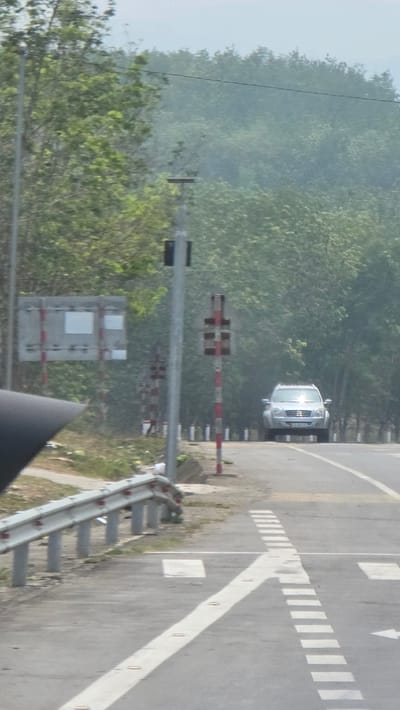 The image depicts a road in Đăk La, Vietnam. A silver car is on the road, approaching what appears to be a railroad crossing, indicated by striped posts and signage. The road has painted markings including a directional arrow and dashed lines. A metal guardrail is visible on the roadside. The environment includes trees and greenery in the background and on the side of the road, with a misty atmosphere.
