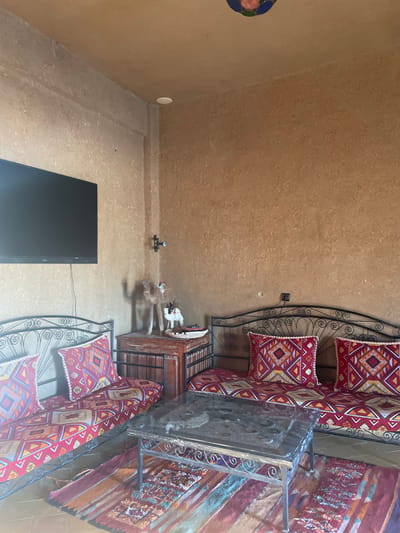 A room with brown walls and ceiling, furnished with two metal-framed seating areas with colorful cushions and a matching rug. Between them sits a wooden cabinet and a glass-topped table. A black rectangular object rests on the floor near the bottom left corner of the room. On the wall, there is a small circular decoration and a small object near the center.