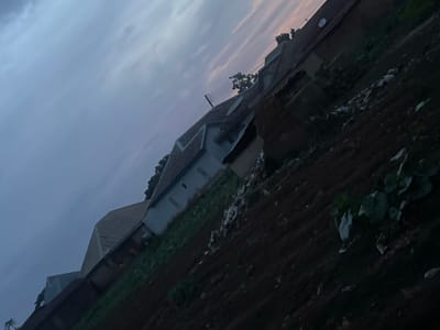 The image depicts a dimly lit outdoor scene, angled with the sky dominating the upper left portion. The sky exhibits soft, horizontal bands of grey and pale pink, suggesting either dawn or dusk. Below the sky, a series of buildings with tiled roofs are visible. The structures are of varying heights and designs, with some appearing to be residential homes. The foreground and midground consist of dark, tilled earth, possibly a garden or field. Scattered across the dark earth are some lighter-colored debris, including what appear to be white plastic bags and other refuse. Some green vegetation, possibly crops, is also present in the lower right section of the image, with large, dark green leaves. The overall impression is of a rural or semi-urban environment in Kassa, Nigeria, at a time of low light. There are no people visible in the frame.