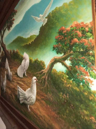 The painting depicts a tranquil landscape with doves. In the sky, a single dove soars. On a gentle slope, a flock of white doves is scattered among lush green bushes and a tree with vibrant red flowers. The background features rolling green hills under a partly cloudy blue sky. The painting is framed with dark wood.