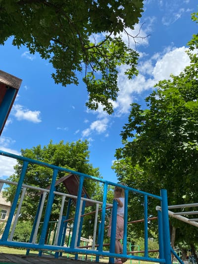 In Markivka, Ukraine, a young child with blonde hair, wearing a white t-shirt and pink shorts, stands on a blue and white playground structure. The child faces away from the camera, looking towards the right side of the frame. The playground equipment appears to be made of metal pipes and wooden planks. In the background, lush green trees are abundant, with their leaves reaching towards a bright blue sky filled with scattered white clouds. To the left, a portion of a brick building is visible. The overall scene suggests an outdoor recreational area on a sunny day.