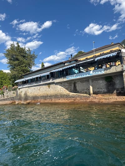 This image depicts a waterside establishment, likely a restaurant or cafe, located along the shore of Lake Maggiore in Magadino, Switzerland. The building features a light blue facade with striped blue and white awnings extended over a terrace. Below the terrace, the building is supported by a stone wall that descends to the water's edge. The water is a deep teal color, with small waves and some scattered white foam. A large green tree stands to the left of the building, and further greenery and flowers are visible behind the stone wall. The sky is a vibrant blue with scattered white cumulus clouds. A small white banner with text is visible on the underside of the terrace. The scene is well-lit, suggesting it was taken on a sunny day. There are no people visible in the frame.