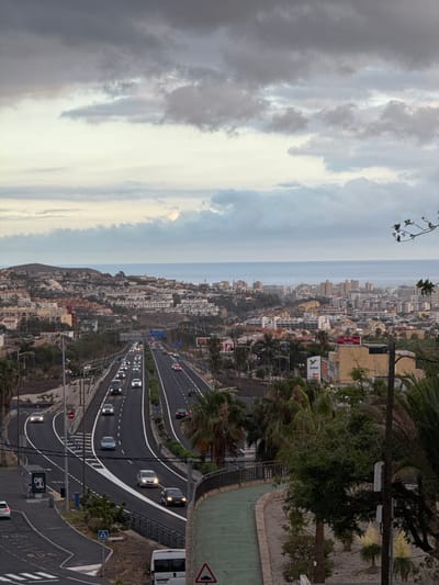 The image shows an elevated view of a coastal city with a highway running through it, near Fañabé, Spain. The city, characterized by its densely packed buildings with white and light-colored facades, sprawls up a hillside towards the sea. The sea is visible in the distance, meeting the horizon under a cloudy sky.

The highway is a prominent feature, with multiple lanes curving through the landscape. Several cars are visible on the highway, their headlights on, indicating it might be dusk or early evening. The weather appears overcast, with heavy clouds dominating the sky, though some lighter areas suggest the sun may be trying to break through. Palm trees and other lush vegetation are scattered throughout the scene, especially on the hillsides and along the edges of the highway, hinting at a warm climate.

In the foreground, the highway is bordered by concrete barriers and some landscaping. A green paved area is visible to the right of the highway, potentially a walkway or recreational space. A white bus is parked on the side of the highway, and a pedestrian crossing sign is visible.

Some text is visible on signs along the highway and on buildings in the city. One sign on a building in the distance appears to be an advertisement for Coca-Cola. Another sign on a traffic light pole reads "CHC". There is also a triangular road sign with a black symbol on a white background.

The overall impression is a busy urban environment situated in a scenic coastal location, with the activity of traffic continuing into the evening under a dramatic sky.