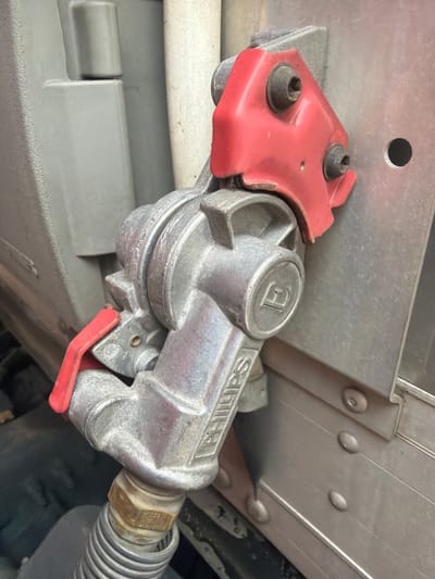 The image is a close-up, low-angle shot of a refueling nozzle with a red lever, attached to a metal conduit. The nozzle is silver and has the word "PHILIPS" embossed on it, along with a circular emblem containing the letter "E." The nozzle is connected to a corrugated hose at the bottom. Behind the nozzle, a section of a gray, textured surface is visible, possibly part of a vehicle. To the right of the nozzle, a silver metal panel with horizontal ridges and several circular rivets is present, consistent with the exterior of a truck or trailer. A small, circular dark hole is visible in this panel. The overall scene appears to be outdoors, possibly in an industrial or service area, but no specific location in Murfreesboro, United States of America, can be determined from the image alone. There are no people or discernible actions occurring in the frame.