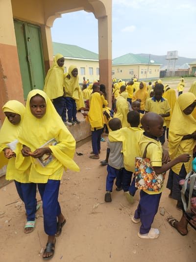 In Kwadon, Nigeria, a group of children in yellow hijabs and uniforms are gathered outside a building. The building has light yellow walls, a green door, and an arched entrance. Several children are in the foreground, some holding books, others looking around. In the background, more children are visible, along with more buildings and a water tower. The ground is dusty, and the sky is overcast. The overall scene appears to be a school setting.