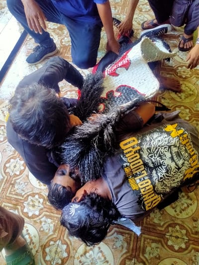 The image depicts an indoor scene in Menjing, Indonesia, on a light-colored ceramic tile floor with repeating geometric and floral patterns in brown and beige. Five individuals are visible.
In the foreground, two adult males are lying supine on the floor with their heads in close proximity. The male on the right wears a dark gray t-shirt featuring a prominent yellow and white tiger graphic and text; a black and white patterned bandana covers his forehead. His face, and that of the adjacent male in a black shirt, exhibit dark smudges or markings. Both appear to be in a state of unresponsiveness or trance. A third individual, wearing a black long-sleeved shirt and an extensive amount of shaggy black material around the neck and upper torso (possibly part of a costume), is lying prone over the two supine males, their head positioned over and pressing against them.
At the upper left, a fourth individual is kneeling, wearing a blue t-shirt, dark blue pants, and black sneakers. Their hands are placed on a large, segmented costume element. This object, primarily white with red and black stylized patterns, appears to be constructed from woven or rigid material, resembling a component of a traditional headdress or animal mask. To the upper right, a fifth individual is partially visible, wearing dark pants and dark flip-flops, with their hands also on the aforementioned costume element. Additional hands are visible around the costume, and a foot wearing a green flip-flop is visible in the lower left corner.
The interaction suggests a structured event, possibly a traditional cultural performance or ritual, where three individuals are in a state of altered consciousness or performance, with others actively attending to them and manipulating a prominent costume piece.