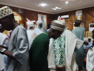 The image shows a group of men in a room in Kaltungo, Nigeria, located at coordinates 9.816 latitude and 11.315 longitude. The men are all wearing traditional clothing, some with white robes and others with green and white robes. They are standing in a group, some with their hands raised, appearing to be in a state of prayer or in a celebratory gathering. The room is decorated with wooden paneling and has a few framed portraits on the walls. The ceiling has recessed lighting. The men are positioned in front of a partially visible doorway and a window, with a framed portrait of a man in a white robe and a hat hanging on the wall in the background. The focus is on the man in the center of the image, wearing a white robe with a green patterned vest, who is being surrounded by other men.  There is a man on the left edge of the image who is partially facing the camera, looking away from the group with his hands outstretched as he speaks to the group.  There is a doorway with an arch on the left side of the room, and a wall with wooden panels and a framed portrait above the doorway.  The image is slightly blurry, suggesting it was taken quickly or perhaps there is a slight motion blur. The image appears to be taken from the perspective of a witness of the event, capturing a moment of  religious ritual or a ceremonial celebration.