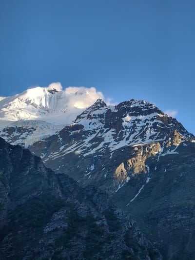This media file is a vertical, eye-level, full shot of a majestic mountain range under a clear blue sky. The primary subjects are the rugged, snow-capped peaks that dominate the frame. The mountain on the left is largely covered in bright white snow and features a prominent glacier. The mountain in the center and right is darker, with patches of snow clinging to its rocky slopes, illuminated by dramatic, warm sunlight, suggesting either sunrise or sunset. Wisps of clouds are present, particularly around the highest snow-covered peak. The foreground features darker, less snow-covered rocky slopes, with some sparse vegetation visible in the shadowed areas.
The scene conveys a sense of awe and the grandeur of nature. The lighting indicates a specific time of day, casting long shadows and highlighting the textures of the mountains. The weather appears clear, with the exception of a few clouds. The location cues point to a high-altitude mountainous region. No people or discernible objects are visible, and there is no visible text. The overall emotion evoked is one of natural beauty and immense scale.