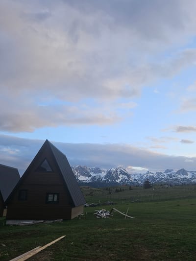 A landscape-oriented image depicts a wooden A-frame house with a black roof, set against a backdrop of mountains with snow-covered peaks and a partly cloudy sky. The house has a triangular window near the roof peak and a rectangular window below. The house sits on green grass. In the distance, there is a scene that could be in the city of Palež, Montenegro. The sky is a mix of blue and grey clouds, and the overall scene seems to be outdoors during daytime. There is lumber next to the house.