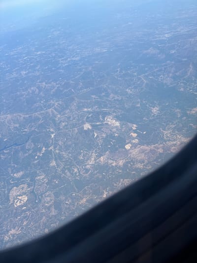 This is a high-altitude aerial photograph captured through an aircraft window, depicting a wide expanse of rural landscape within the region of Saint-Georges-d'Espéranche, France. The terrain is characterized by undulating hills and valleys covered by a mosaic of dark green vegetation, likely forests, intermingled with lighter tan and brown areas indicating fields, clearings, and potentially sparse ground cover. A prominent, sinuous river or large stream traverses the scene, appearing as a dark, winding ribbon. Numerous fine linear elements, consistent with roads or agricultural boundaries, are observable across the topography. Several irregular, bright patches suggest large-scale ground disturbances, possibly quarries or extensive construction sites. The upper portion of the image shows a hazy light blue sky. The lower right quadrant is partially obscured by the dark, curved frame of the aircraft window. No individuals, specific actions, or discernible structures are visible due to the extreme altitude and distance. The environment is uniformly illuminated, suggesting clear daylight conditions.