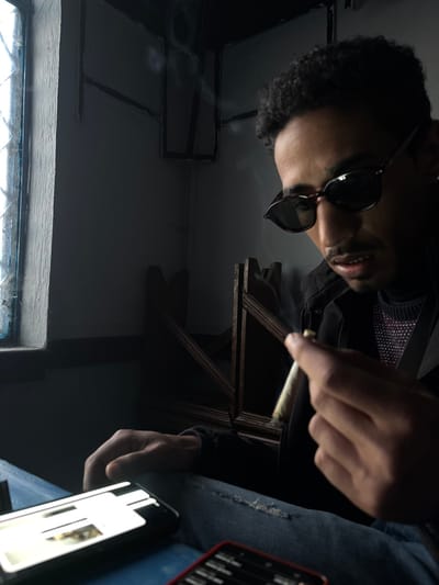 An upward shot features a man with curly hair and sunglasses holding a lit cigarette. He wears a dark jacket over a patterned top and blue jeans. A smartphone lies nearby. The backdrop includes a window with gridded panes and a dark wall. A wooden structure partially obscures the wall, with a faint smoke trail visible near it. Lighting is dim, creating a somewhat shadowy ambiance.