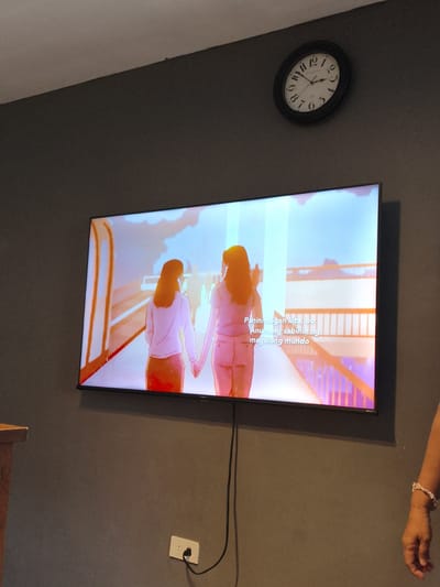 The image shows a flat-screen television mounted on a dark gray wall. On the television, an animated scene depicts two young women walking hand-in-hand, with subtitles in a foreign language displayed at the bottom right. Above the television, a round analog clock with black numbers and hands is mounted on the wall. To the left of the television, a small portion of a wooden table is visible. Below the television, an electrical outlet with a plug and black cable is attached to the wall. A portion of a person's arm with a bracelet is visible on the right side of the image, extending from the bottom edge. The overall environment appears to be indoors, likely a living room. The image does not contain any specific indicators that would allow for a precise location within Cavite City, Philippines.