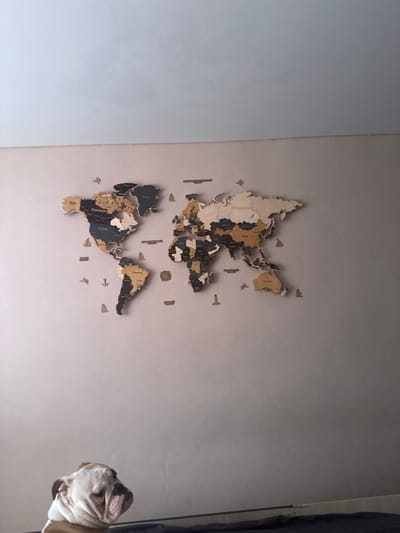 An indoor scene features a wooden world map mounted on a light pink wall. The map is composed of various wooden pieces representing continents and countries, with some labeled in different colors. Small wooden airplanes and sailboats are also attached to the wall around the map. In the lower portion of the image, a bulldog is visible, resting its head and looking downwards. The background above the map appears to be a white ceiling. The overall impression is that of a decorated living space, possibly in a home.