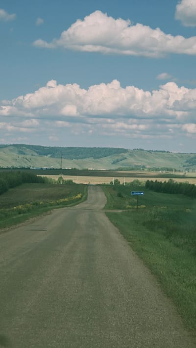 The image shows a rural road in the city of Pobeda, Russia, leading through rolling green hills under a partly cloudy sky. The road is paved and appears to be a two-lane country road. On the right side of the road, a blue sign with white text reads "Д. ЕЛХОВКА →", indicating a direction towards a village. The landscape is characterized by agricultural fields and grassy verges, with a distant line of trees and undulating hills forming the horizon. The overall scene depicts a serene and natural environment.