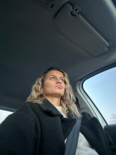 Inside a car, a woman with wavy blonde hair and light eyes gazes upward. She wears a white sweater and a dark, textured coat, secured by a seatbelt. The car's interior features a sun visor and a view of the sky through the windshield. The perspective is from below, looking up at the woman, emphasizing the car's ceiling and window.