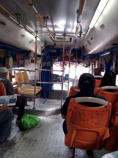 The image shows the interior of a bus in San Lorenzo, Paraguay. There are rows of orange plastic seats occupied by several passengers. A man wearing a baseball cap is seated near the front, reading a paper. A green bag is on the floor near him. The ceiling of the bus is metal and has hanging straps and decorative ornaments. Curtains are around the front of the bus. The floor is a metal diamond tread pattern.