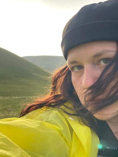 A close-up, selfie-style image shows a person wearing a black beanie and a bright yellow hooded jacket. The person has dark hair that is partially obscuring their face, and a silver nose ring. Their eyes are open and looking towards the camera. The background is a landscape of rolling green hills, suggesting an outdoor, possibly rural or mountainous location. The sky is bright and appears to be either sunrise or sunset, with the sun creating a strong backlight. The image quality is good, with natural lighting and sharp focus on the person's face. There are no visible elements that specifically indicate the location is the city of Donskoe, Russia.