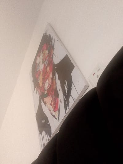 The media file captures an indoor scene, likely in Abeokuta, Nigeria, featuring a brightly colored, abstract painting mounted on a light-colored wall. The painting, possibly an expressionistic portrait, is characterized by broad, impasto-like strokes of red, white, and yellow, contrasted with heavy black brushwork and prominent black drips.

The image is captured from a low, tilted angle, giving a diagonal perspective of the wall and the artwork. To the right of the painting, a white electrical outlet or switch with a small red indicator light is visible on the same wall. In the bottom right corner and along the right edge, a dark, upholstered object, possibly a part of a chair or couch, partially frames the scene.

The lighting is soft and ambient, preventing determination of the specific time of day. There are no people visible, and no specific activity or event is taking place. The overall mood is quiet and still. No clearly legible text is present on the painting or elsewhere in the image. The photograph exhibits a slight grainy texture and a warm, muted color cast.