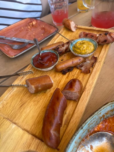 In Byurakan, Armenia, a wooden board filled with sausages and various accompaniments is presented on a table. The board holds several cooked sausages, some whole and some sliced, along with pieces of crispy bacon. Two small bowls are also on the board: one contains a dollop of mustard, and the other holds ketchup. Serving tongs are positioned to pick up a sausage slice. To the left of the board, a partially eaten meal is visible on an orange rectangular plate, with a fork and knife resting on it. In the background, a person's arm wearing a horizontally striped shirt is visible, along with glasses and pitchers of red and yellow beverages. A blue-rimmed bowl containing a red sauce with a spoon is also partially visible in the lower right corner of the image. The overall scene depicts a casual meal, likely outdoors, given the wooden table and natural lighting.