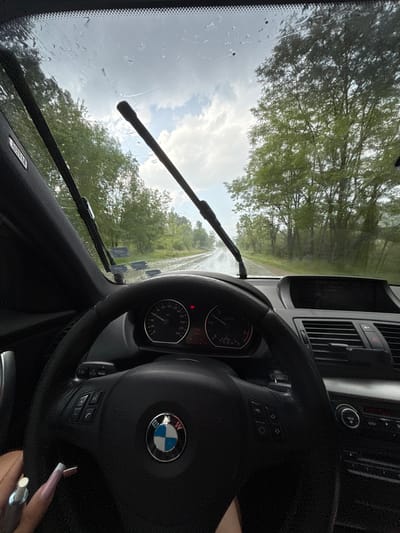 The media file captures an interior view from a car in Kaleytsa, Bulgaria, looking out through the front windshield. The scene depicts a rainy or recently rained-on day, with raindrops and water streaks covering the glass and the car's wipers visible.
The subjects include the car's interior, specifically the steering wheel and dashboard, and the exterior environment. The car is identifiable as a BMW by the emblem on the steering wheel. The car appears to be stationary, as the speedometer needle rests at zero. Outside, a road or path is visible, flanked by numerous green trees, suggesting a natural or possibly rural setting. The sky is overcast with patches of brighter clouds.
Notable details include the time of day, which is daytime. The dashboard features a speedometer (marked up to 240 km/h) and a tachometer, along with a digital display showing "18:92," "-19.5," and "99.9%." A "MAX" button is visible on the central console, and "AIRBAG" text is printed on the car's pillar. Part of the driver's right hand is visible in the lower right corner, adorned with long, manicured nails (French tip style) and a silver ring on the ring finger, holding a small white object.
