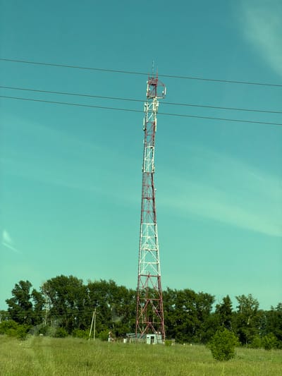 A red and white lattice tower, likely a cell tower, stands tall in a grassy field with a line of green trees behind it. Three black wires stretch horizontally across the top of the frame, above the tower. The sky is a clear, bright teal with faint wispy clouds. At the base of the tower, there is a small white building.