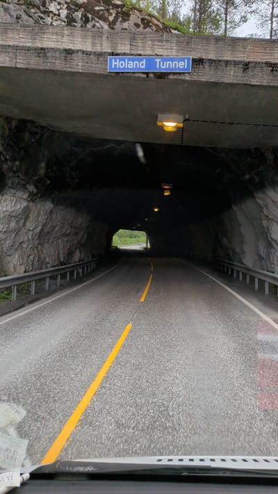 The image shows the entrance to the Holand Tunnel in Mosjøen, Norway. A blue sign with white lettering reading "Holand Tunnel" is mounted on the concrete structure above the tunnel entrance. The tunnel is carved into a rocky mountainside, with rough, grey rock formations visible on the sides. A metal guardrail runs along the left side of the road leading into the tunnel. The road surface is asphalt with a prominent yellow line marking the center. Several yellow lights are visible inside the dark tunnel, illuminating the path. The exit of the tunnel, visible in the distance, shows daylight and a hint of greenery, suggesting an outdoor environment beyond the tunnel. The photo is taken from a vehicle's perspective, looking straight ahead into the tunnel.