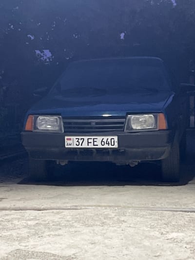 The image shows the front of a dark blue car parked on a paved surface. The car has its headlights on and its license plate is visible. The license plate reads "AM 37 FE 640". The car appears to be in a garage or covered area, as indicated by the dark shadows and overhead foliage. The lighting is dim, suggesting it might be dusk or night. The overall environment is urban. The car is the primary subject of the image.