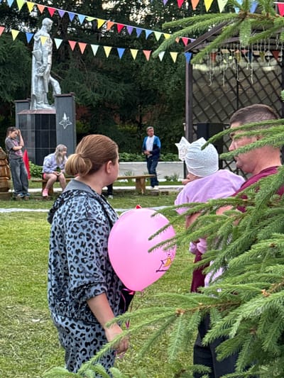 In the city of Kukui, Russia, an outdoor gathering is taking place. A woman with her hair in a bun, wearing a grey leopard-print jacket, holds a pink balloon with a cartoon character on it. She stands near a man in a dark red shirt, who is holding a baby wearing a white bow and a pink outfit. In the background, a young girl with blonde hair sits on a wooden bench, and another girl in a grey t-shirt and jeans stands nearby, resting her hand on her head. A monument with a statue is visible behind them, adorned with colorful triangular flags strung above. A gazebo with decorative elements and hanging lights is also present. The ground is covered in green grass, and a large spruce tree partially obscures the foreground.