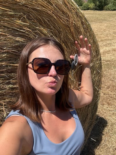 A woman with brown hair and sunglasses poses in front of a large hay bale in a dry, grassy field. She is wearing a blue tank top and a silver necklace. Her right hand is raised, holding a set of keys. The sun is shining, casting shadows on the hay bale and the ground. The background shows more fields and trees, suggesting a rural location, likely in the vicinity of Coteaux du Lizon, France, given the agricultural landscape.
