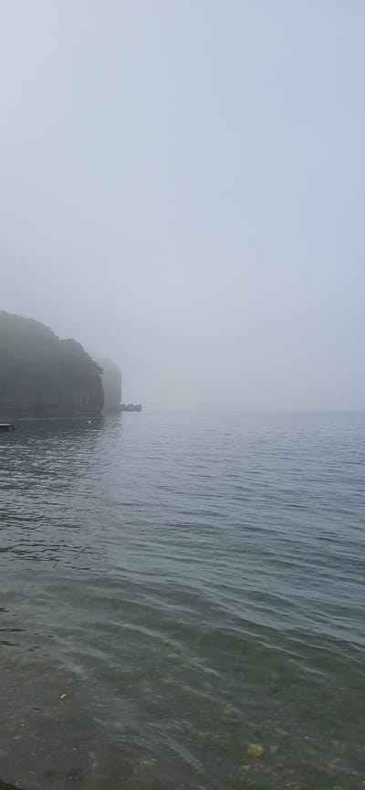 The image depicts a foggy seascape in Slavyanka, Russia. The foreground shows clear, shallow water with visible stones and debris on the seabed. In the mid-ground, calm, gray water stretches out towards a rocky, tree-covered cliff face partially obscured by fog. Several large, irregular rocks are visible in the water near the base of the cliff. A small, white buoy floats on the water to the left of the cliff. The background is dominated by thick fog, creating a hazy, indistinct horizon where the sea meets the sky. No people or specific actions are discernible in the scene.