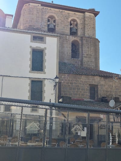 A sunlit urban scene in Piedrahíta, Spain, features architectural elements and a commercial establishment under a clear blue sky. Dominating the background is a multi-tiered stone church tower, constructed from light brown blocks. It displays three visible arched bell openings, each containing a large metal bell, with the upper two featuring decorative supports. A bird's nest is visible on the tower's upper right cornice, and small birds perch along its roofline.
To the left of the tower, a two-story building with a white stucco facade and tan stone trim is prominent. It presents two vertically aligned windows, both fitted with closed dark green louvred shutters. A black street lamp with a single light fixture extends from the facade between the two main windows.
In the foreground, a commercial establishment, identified as "La Cabaña" by white script signage, is enclosed by a clear glass and dark metal barrier. Visible through the glass are multiple empty light-colored wicker-style tables and chairs, some stacked on the right side, indicating an outdoor dining area. Reflections of the surrounding buildings and sky are visible on the glass panels.
To the right of the white building, behind the foreground establishment, another building constructed of stone blocks with a tiled roof and a dark gridded window is partially visible. A small white satellite dish is mounted on its roof. The scene is brightly illuminated by natural sunlight, casting distinct shadows. No individuals are present.