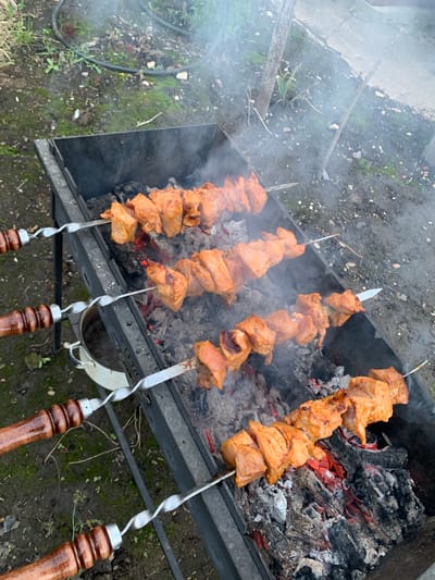 The image depicts four skewers of raw, marinated meat being grilled over hot coals in an outdoor barbecue. The meat appears to be chicken or pork, cut into bite-sized pieces and threaded onto metal skewers. The skewers are placed horizontally across a black metal grill, with some smoke rising from the coals. The environment is an outdoor setting, with dirt, grass, and small rocks visible on the ground surrounding the grill. In the background, a white bucket and what appears to be a hose are present. The lighting suggests an overcast or shaded day. The scene is consistent with a casual outdoor cooking event in a rural or suburban setting. The location is specified as Блеўчыцкі сельскі Савет, Belarus.