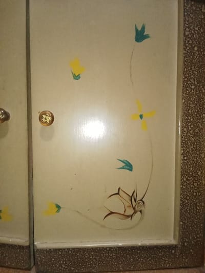 The image displays a close-up, vertical view of two adjacent cabinet or wardrobe doors situated in an interior location in Ciputri, Indonesia. The left door, partially visible, features a round, golden-colored knob with a raised floral or starburst pattern. The right door, which occupies the majority of the frame, is light cream or beige. This door is decorated with hand-painted floral motifs: three distinct yellow flowers with short green stems are located at the upper-left, mid-top, and lower-left sections of the panel. Two teal-colored leaf shapes appear at the upper-right and mid-right. A larger, multi-lobed floral design in shades of brown and lighter beige is positioned in the lower-right corner, from which a thin brown line extends vertically towards the upper-right leaf. A strong, diffuse light reflection is prominent on the central-right portion of the cream-colored door. The bottom edges of both doors and the right edge of the right door are bordered by a dark brown material exhibiting a fine, speckled or granular texture. No individuals or active interactions are present in the scene.